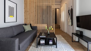 A modern living room with a grey sofa, a black coffee table with books, a wooden partition, and a TV on the right. - Elliot Petralona, Athens (Athens)