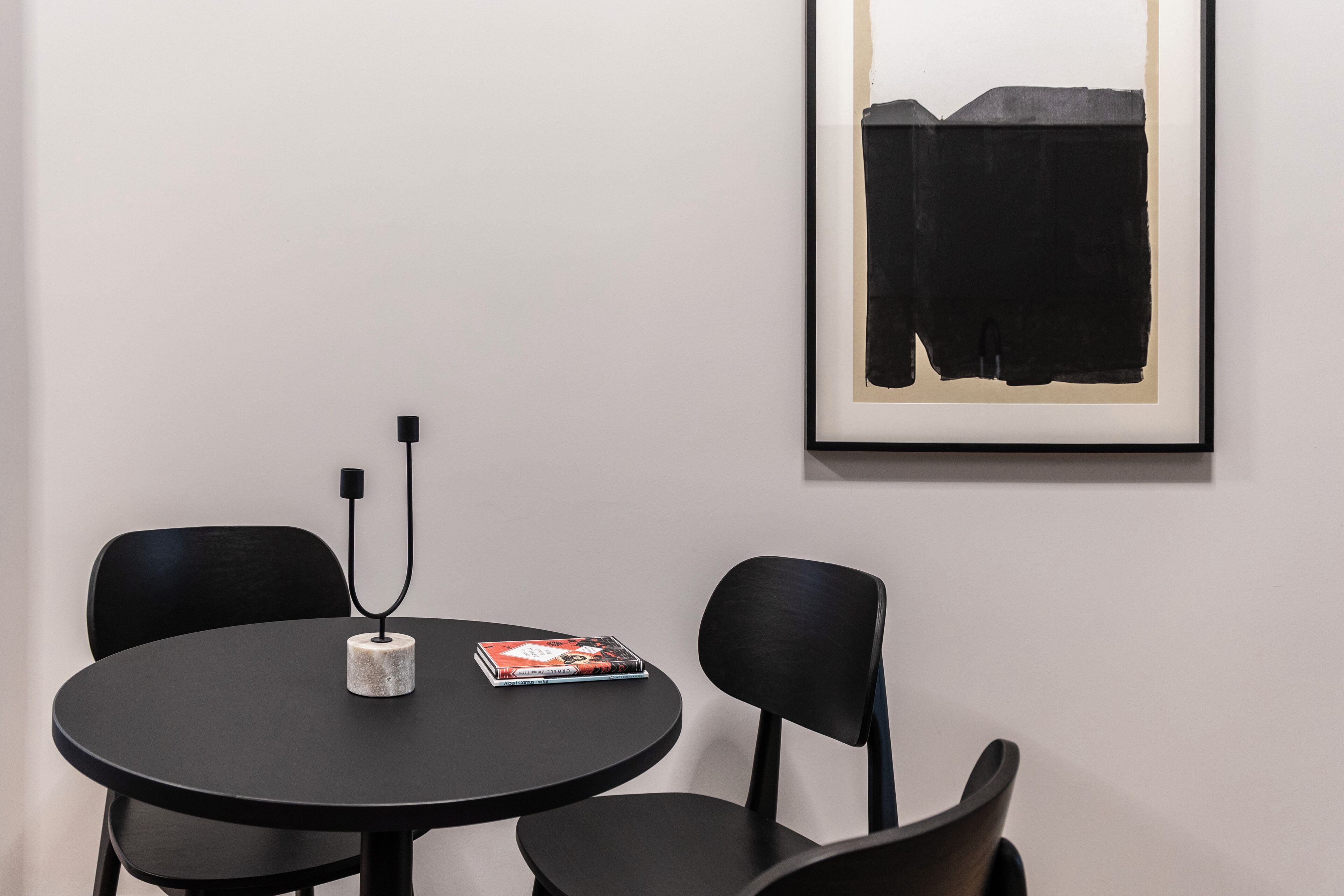 Une table ronde avec un livre et un câble d’alimentation, deux chaises noires et un tableau abstrait encadré accroché au mur.
