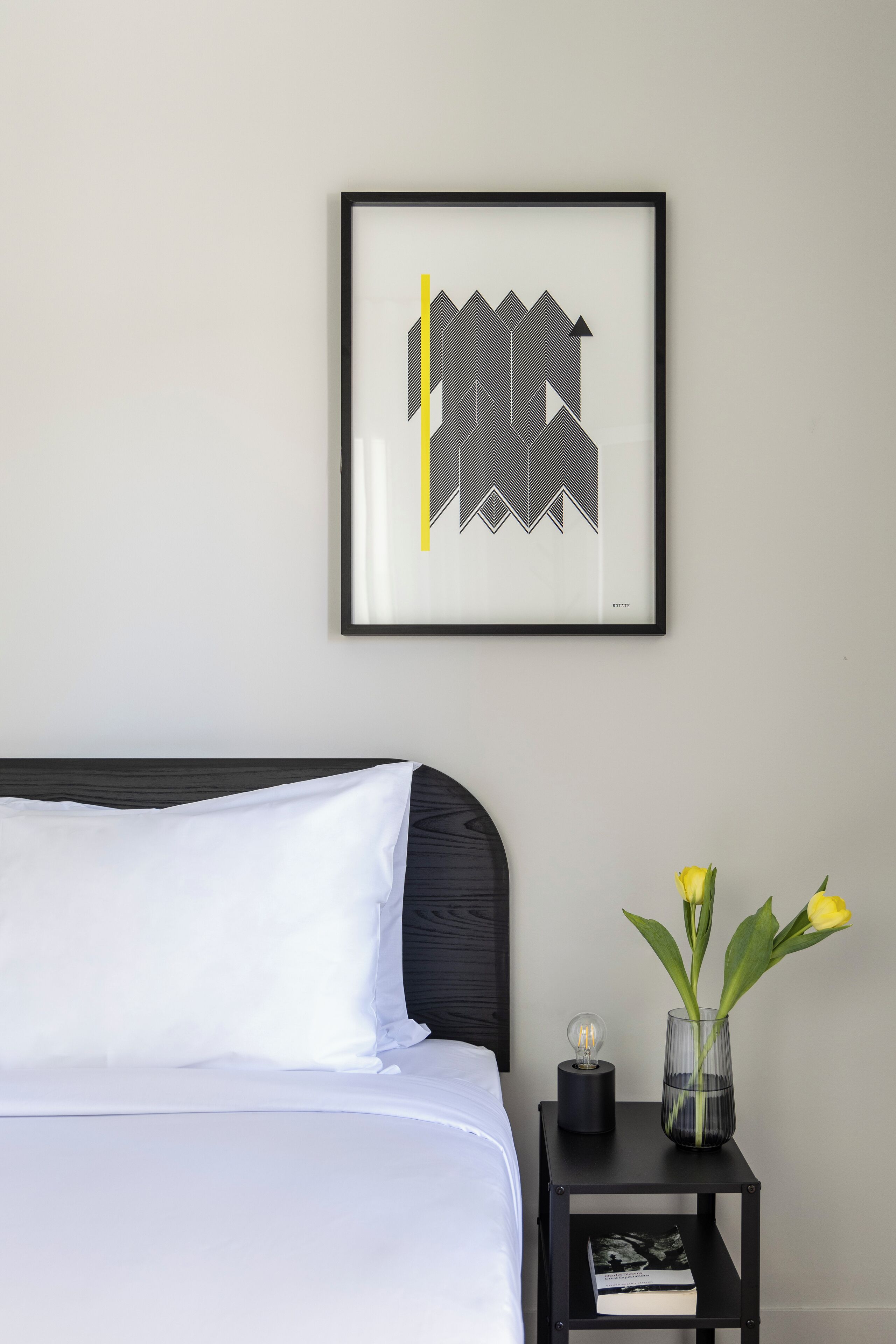 Una cama bien hecha con ropa de cama blanca, un cabecero negro, una obra de arte abstracta enmarcada en la pared, una mesita de noche negra con un jarrón de flores amarillas y un libro.