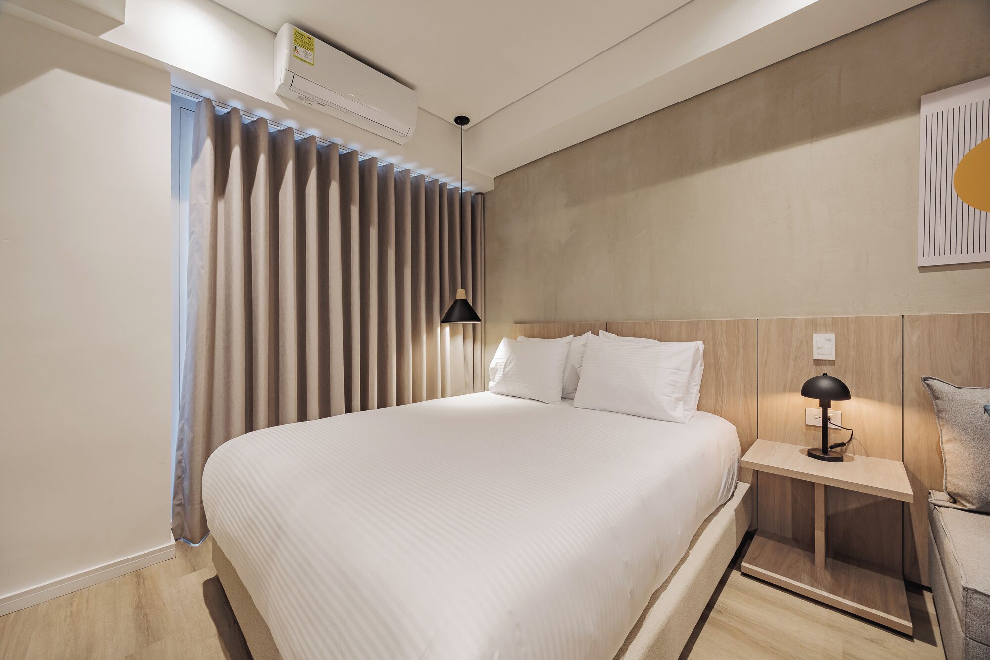 A modern hotel room with a large bed, a bedside table with a lamp, and a wall-mounted air conditioning unit.