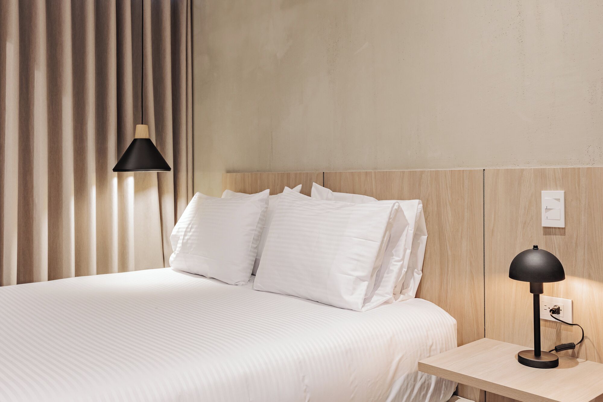 A neatly made bed with white linens, a wooden headboard, a bedside table with a lamp, and a power outlet.