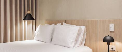 A neatly made bed with white linens, a wooden headboard, a bedside table with a lamp, and a power outlet.