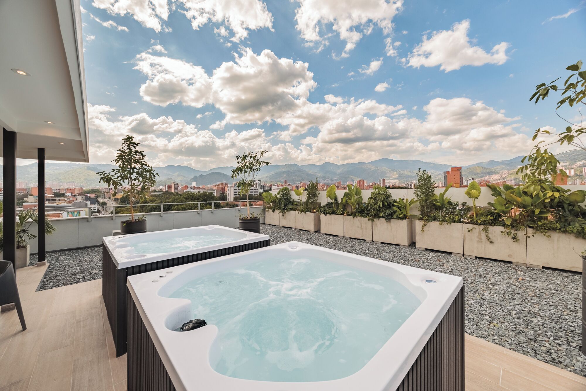 A rooftop terrace with two hot tubs, surrounded by potted plants and a cityscape view.