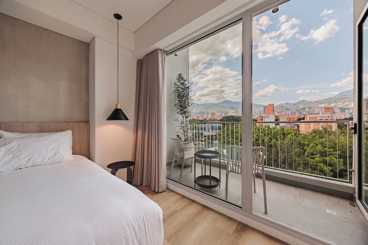 Una habitación de hotel moderna con una cama grande, una mesita y un balcón con vistas a la ciudad y a las montañas.