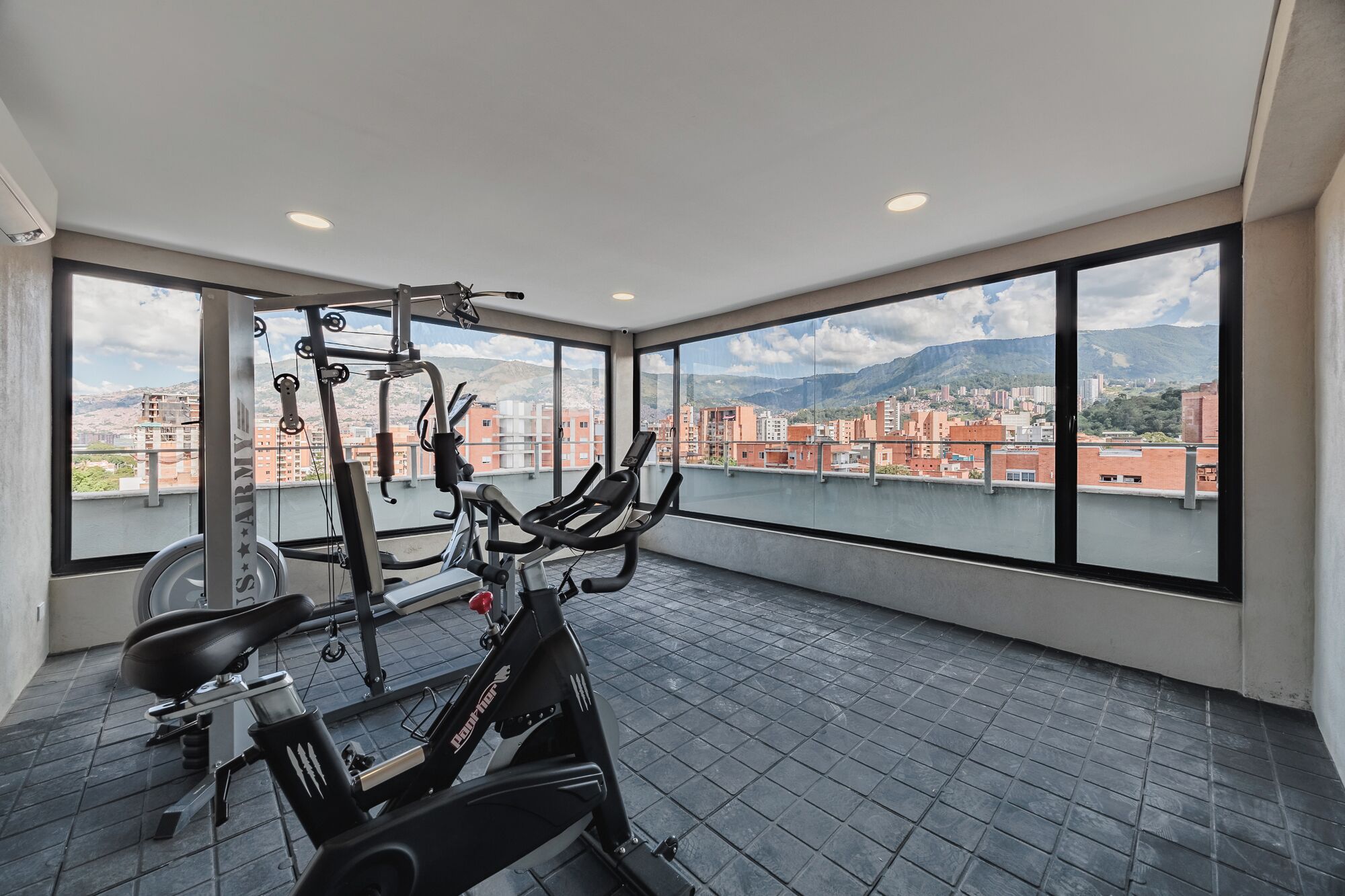 A gym with a stationary bike, a bench, and a weight machine, with a city view through large windows.