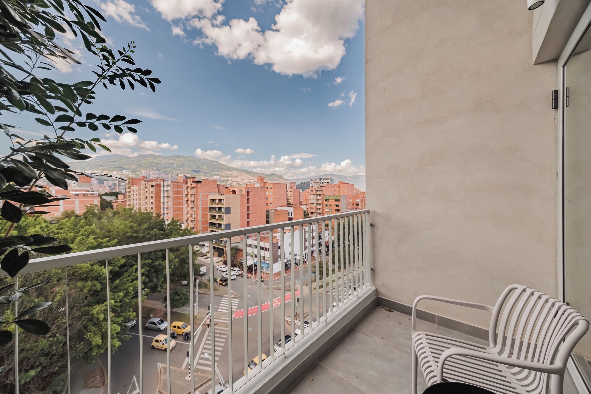 A balcony with a white metal railing, a white metal chair, and a view of a cityscape with buildings and a road with cars.
