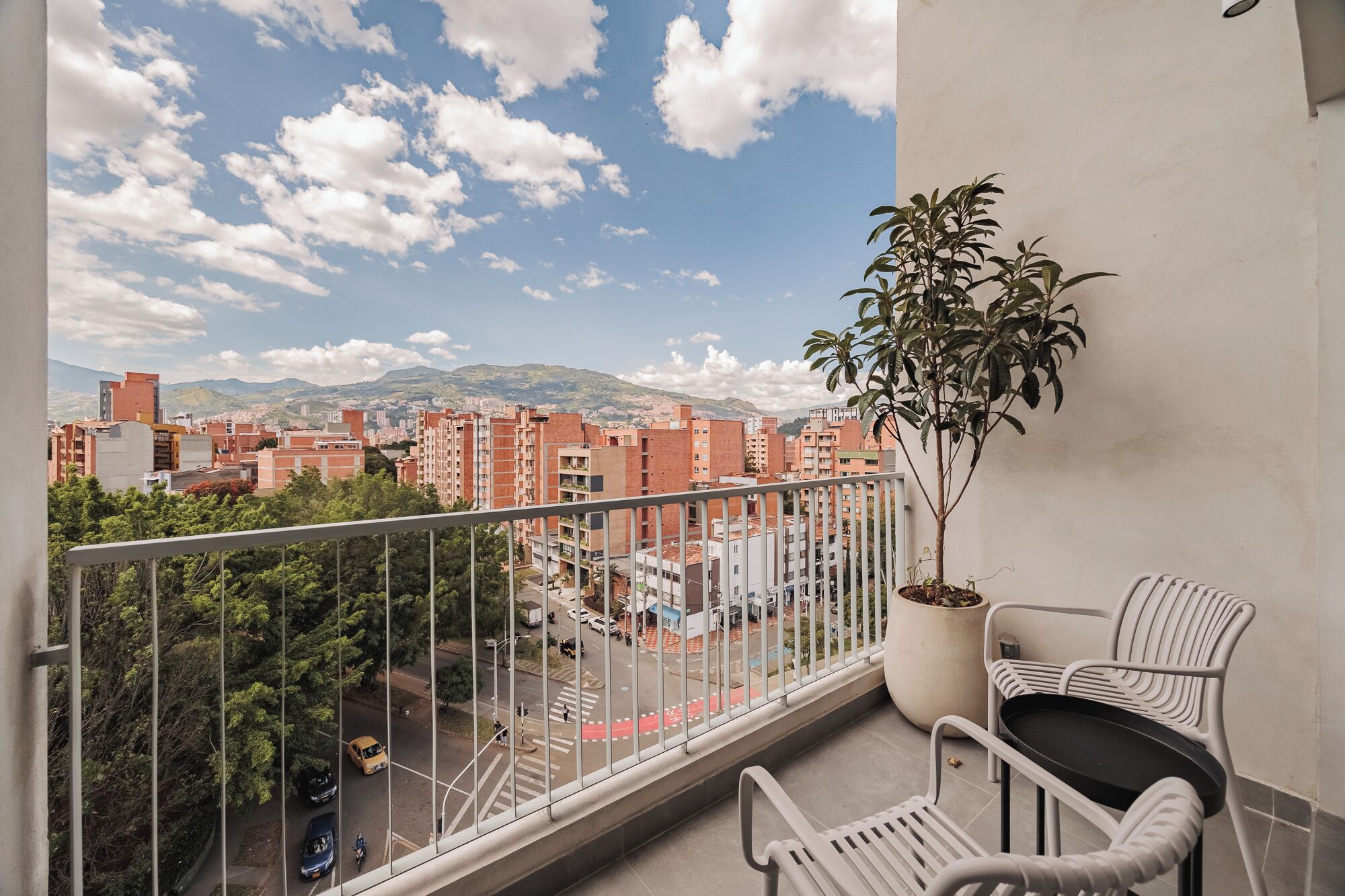 Un balcón con una maceta, dos sillas de mimbre y vistas a un paisaje urbano con edificios y montañas al fondo.