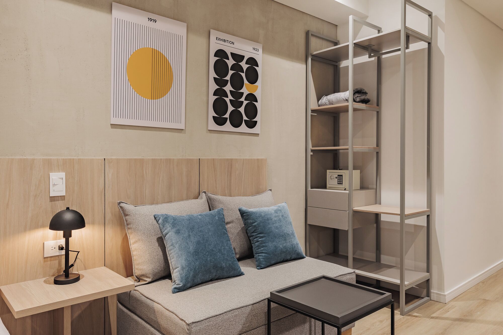 A modern bedroom with a wooden headboard, a bedside table with a lamp, a sofa with blue cushions, and a metal shelving unit.