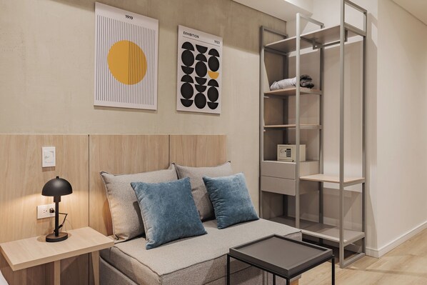 A modern bedroom with a wooden headboard, a bedside table with a lamp, a sofa with blue cushions, and a metal shelving unit.