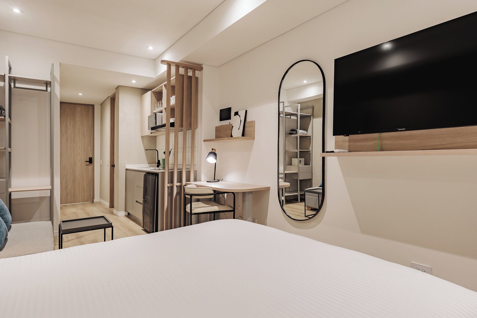 A modern hotel room with a large bed, a desk with a chair, a wardrobe, a mirror, and a TV mounted on the wall.