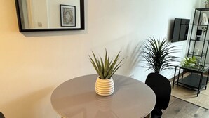 A modern living room with a round table, two black chairs, a potted plant, a framed picture, and a black-framed mirror on the wall. - Cosy One Bedroom London Apartment (London)