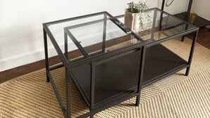 A modern living room with a glass-top coffee table, a flat-screen TV mounted on the wall, and a black metal shelving unit. - Cosy One Bedroom London Apartment (London)