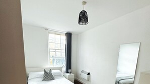 A modern bedroom with a large bed, a bedside table, a heater, and a window with curtains. - Cosy One Bedroom London Apartment (London)