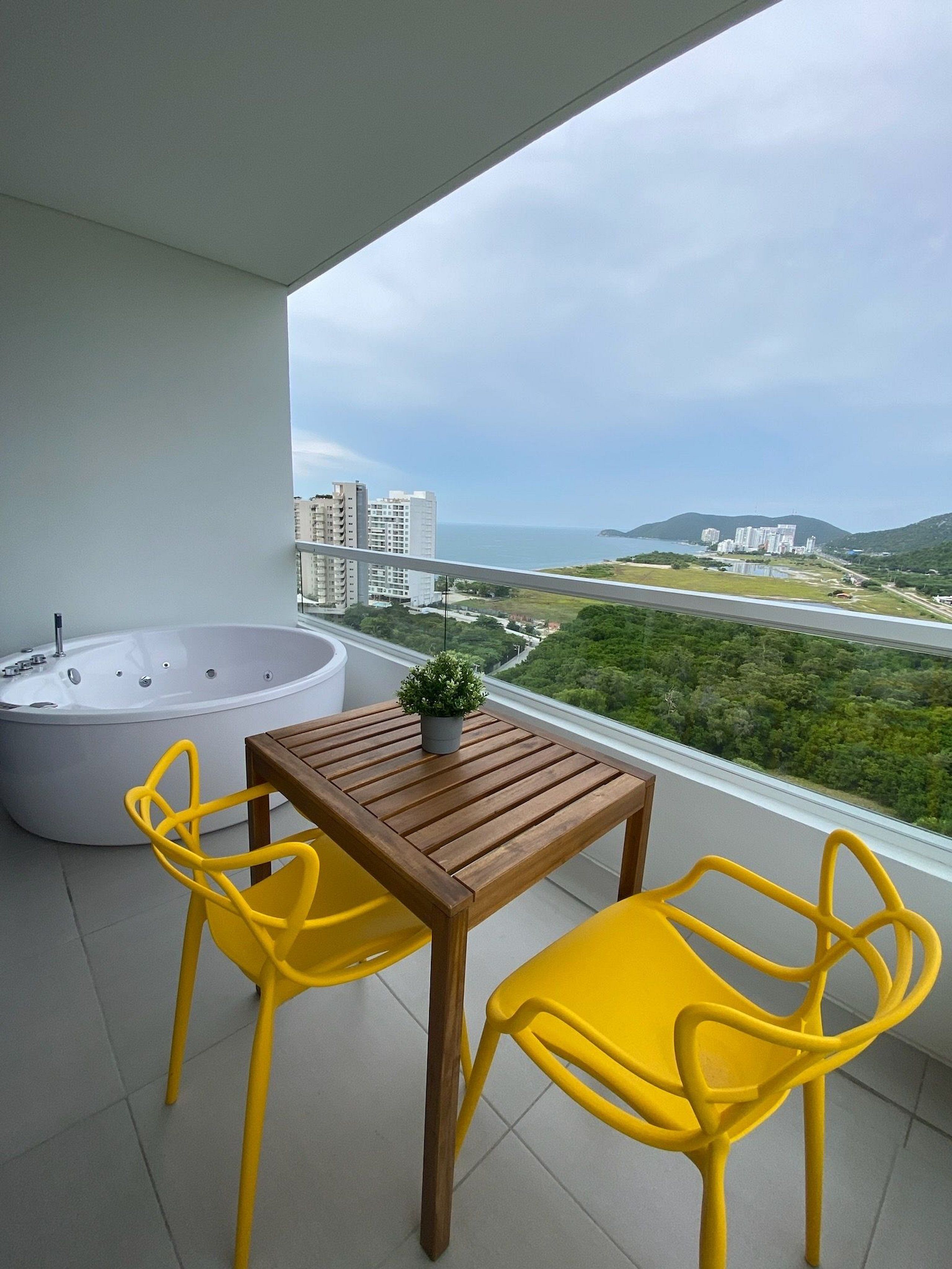 Ein Balkon mit einem Holztisch und zwei gelben Stühlen, einer großen Badewanne sowie Blick auf eine Küstenlandschaft mit Gebäuden und viel Grün.