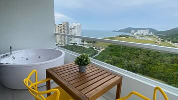 Ein Balkon mit einem Holztisch und zwei gelben Stühlen, einer großen Badewanne sowie Blick auf eine Küstenlandschaft mit Gebäuden und viel Grün.