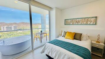 Un dormitorio con una cama grande, un balcón con bañera y vistas a edificios y colinas.