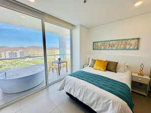Un dormitorio con una cama grande, un balcón con bañera y vistas a edificios y colinas.