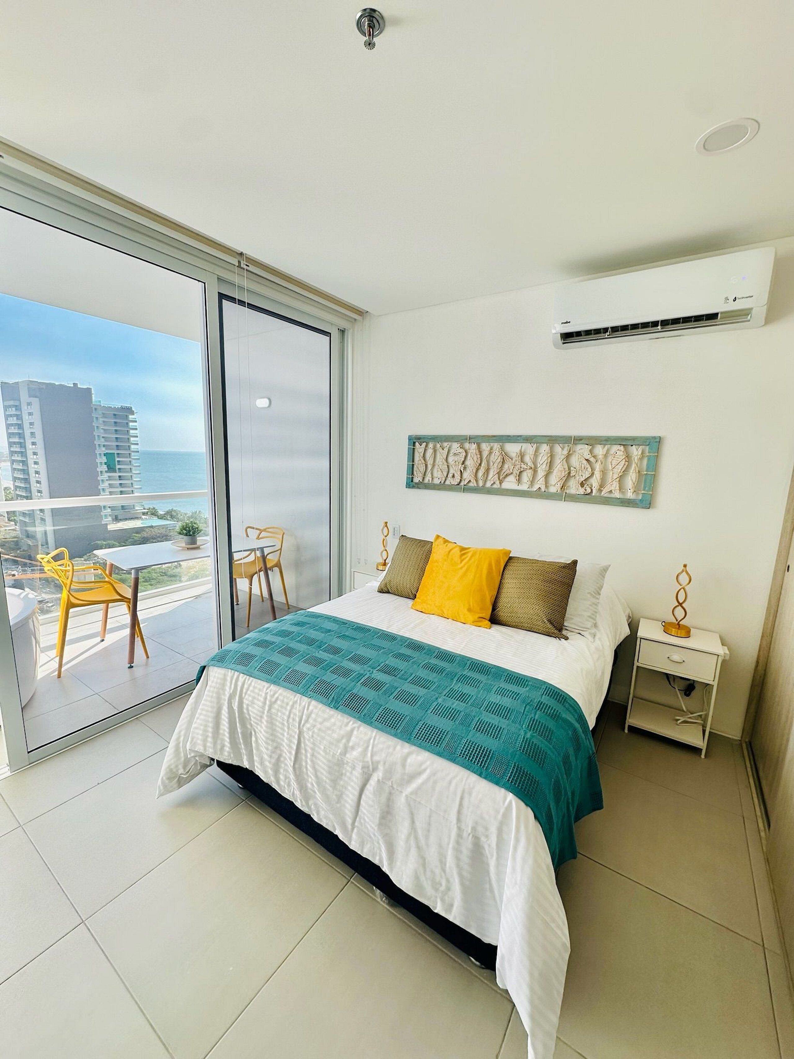 A hotel room with a bed, a nightstand, a wall-mounted air conditioner, and a view of the city through a large window.