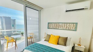 A hotel room with a bed, a nightstand, a wall-mounted air conditioner, and a view of the city through a large window. - U-GO Porto Horizonte Junto al Mar Sta Mta (Santa Marta)