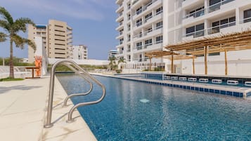 Una piscina con una escalera metálica, rodeada por un edificio blanco con balcones y una palmera.