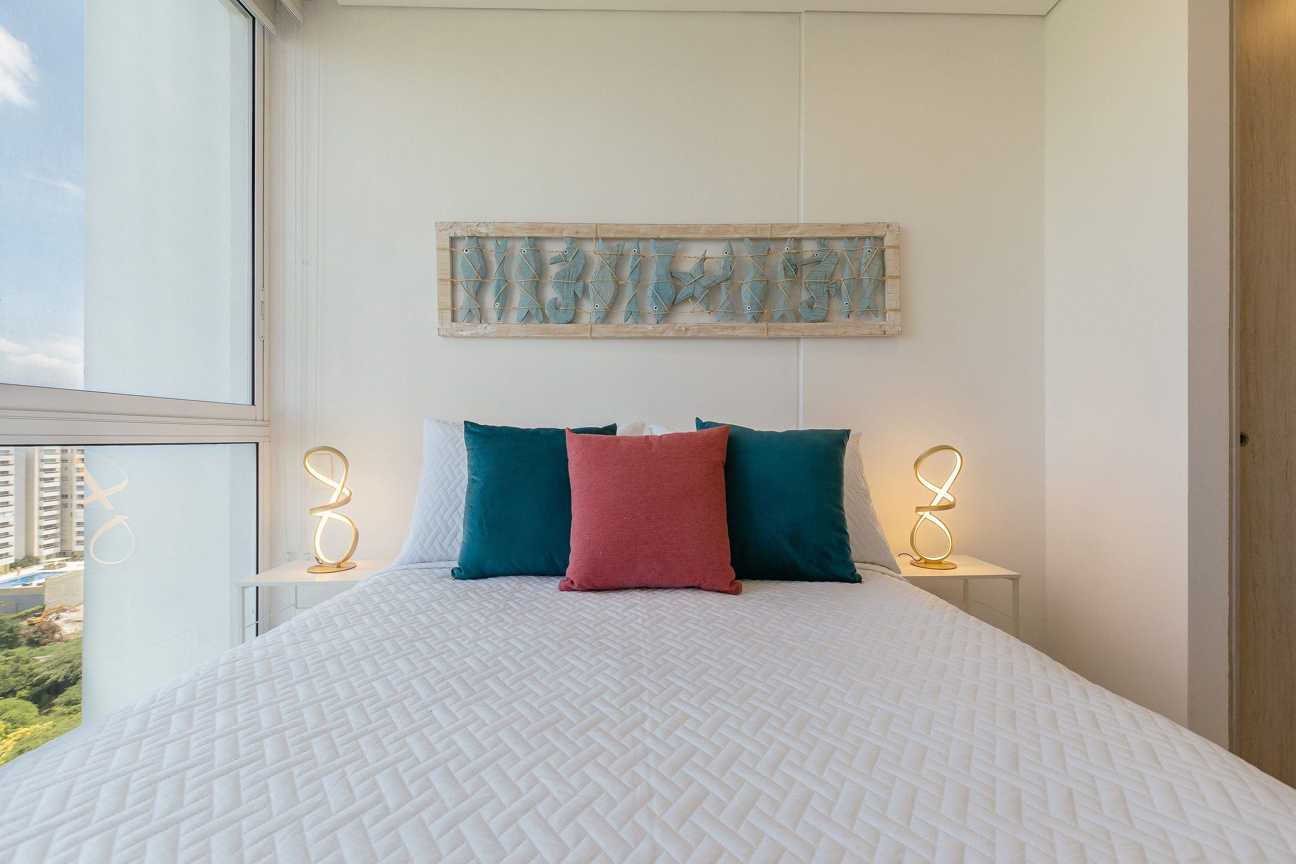 A bedroom with a large bed, two bedside tables with lamps, a wall-mounted artwork, and a view of the outside.