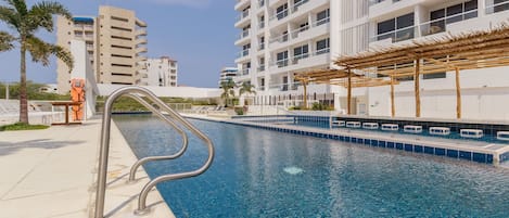 A swimming pool with a metal ladder, surrounded by a white building with balconies and a palm tree.