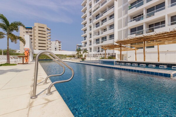 A swimming pool with a metal ladder, surrounded by a white building with balconies and a palm tree.