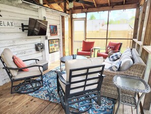 Uma área de estar aconchegante ao ar livre, com móveis de vime, uma TV na parede e um deck de madeira.