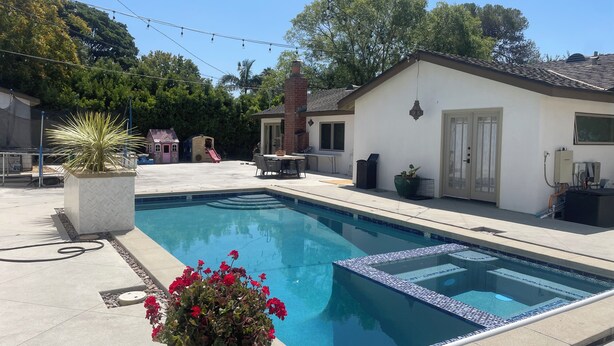 A swimming pool with a tiled edge, surrounded by a patio area with outdoor furniture and a play structure in the background.