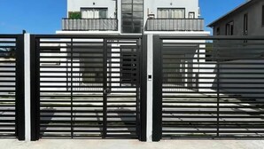 A modern two-story house with a gated entrance, a balcony on the upper floor, and a clear blue sky in the background.