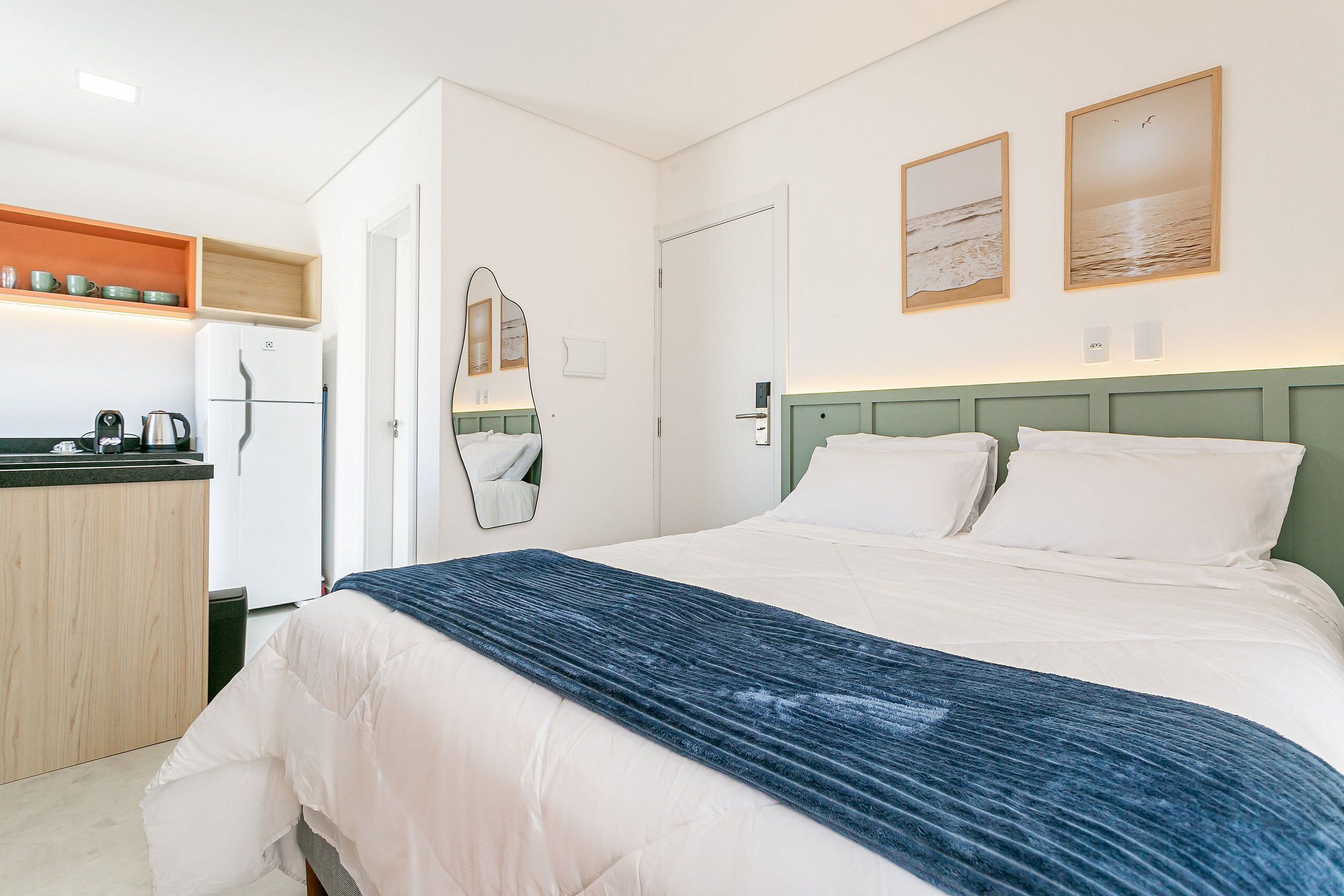 A hotel room with a bed, a small kitchenette, a mirror, and two framed pictures on the wall.
