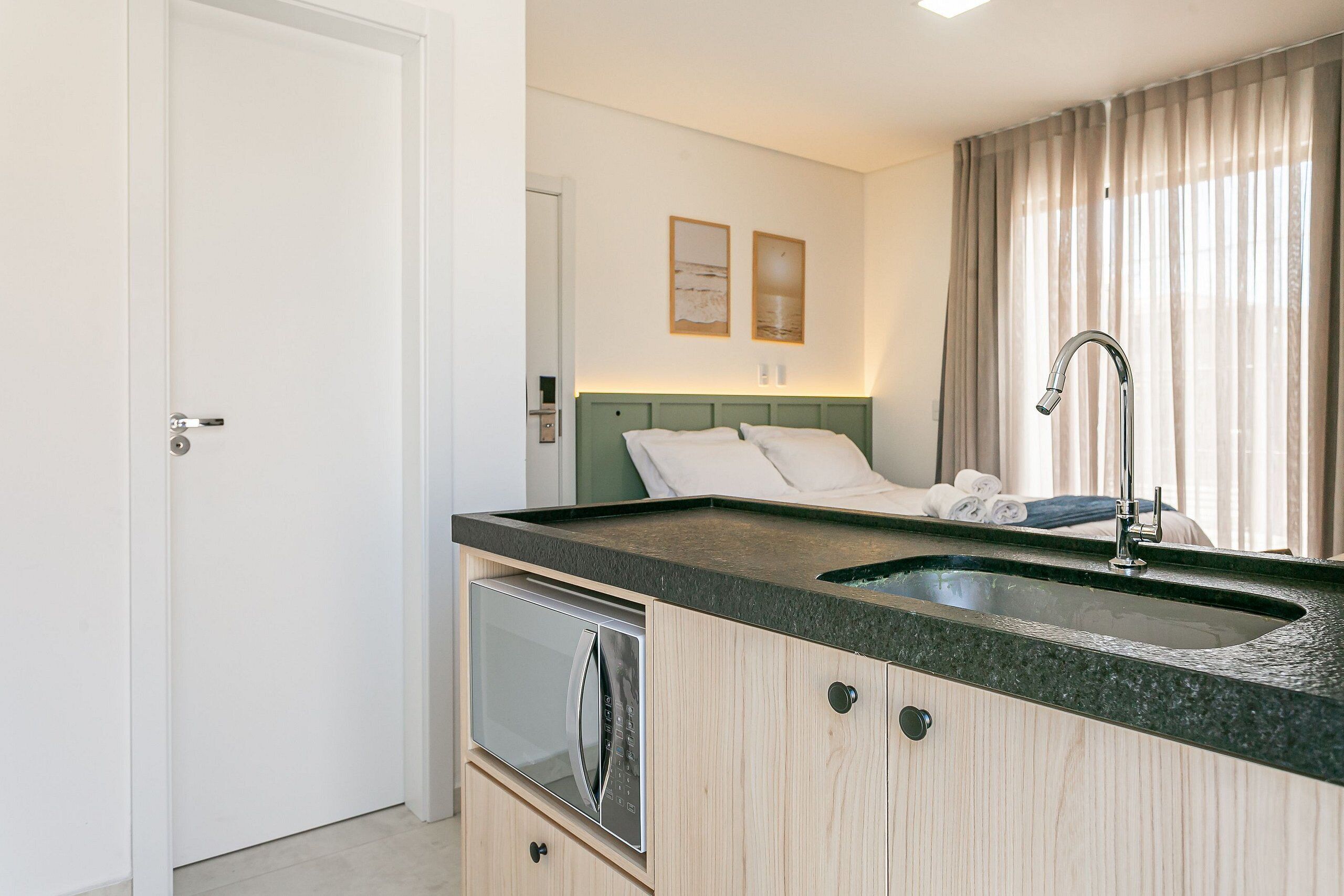 A compact kitchen with a built-in microwave, a sink, and a green headboard in the adjacent bedroom.