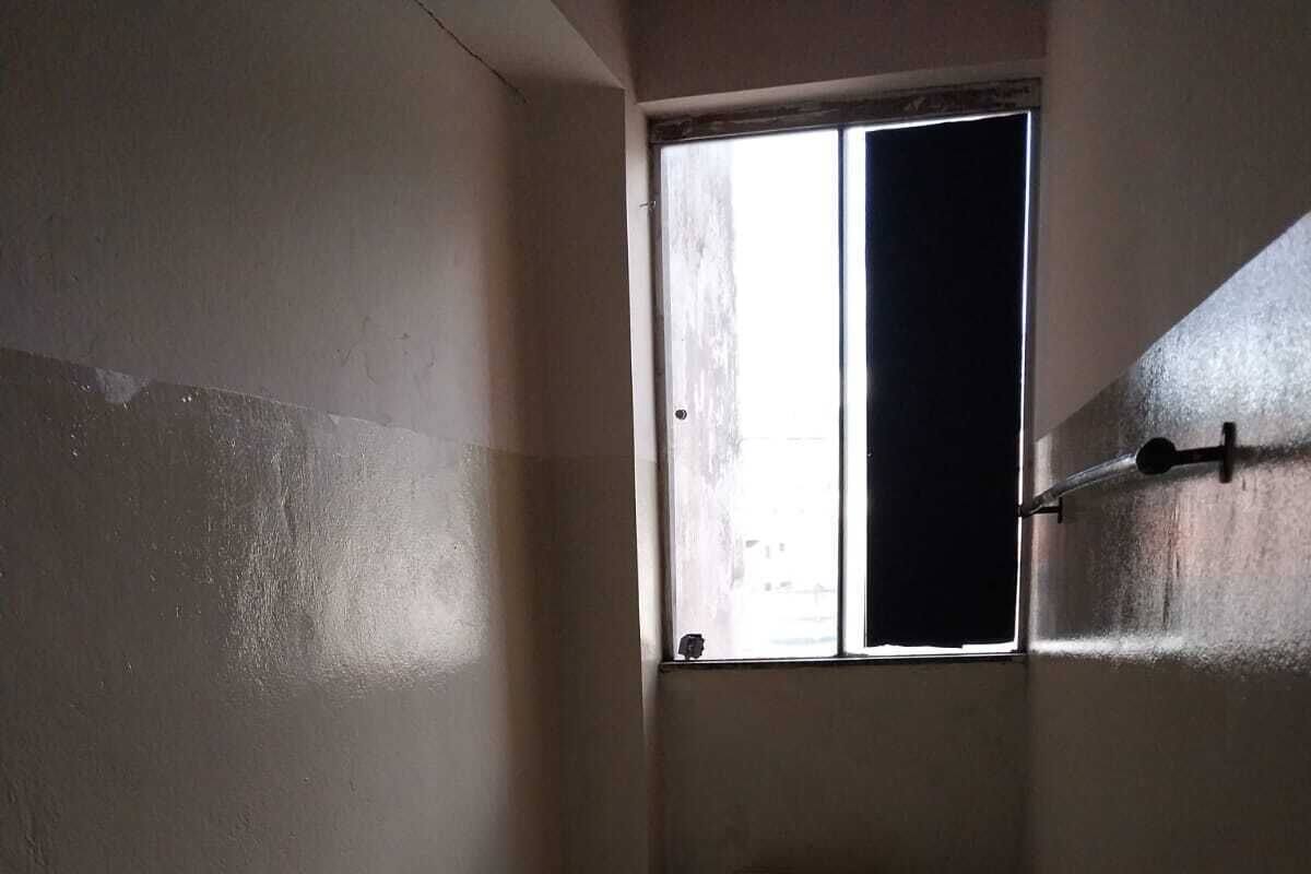 A small, dimly lit room with a single bed, a window, and a metal shelf.