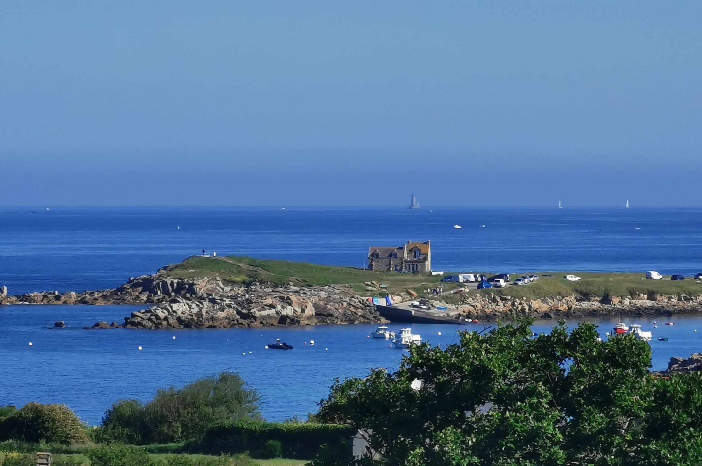 Un paysage côtier avec un bâtiment historique en pierre, situé sur une colline verdoyante, des bateaux sur l’eau et un ciel bleu dégagé.
