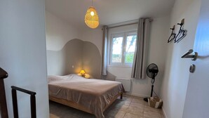 A bedroom with a bed, a bedside lamp, a standing fan, a window with curtains, and a door with a handle. - Pinson, Gîte with outdoor spa ideal for couples (Saint-Avit-Sénieur)