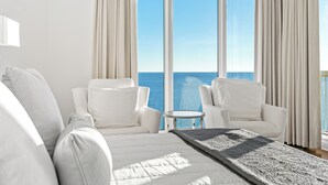 Una habitación de hotel moderna con una cama grande, dos sillones blancos, una mesita redonda y vistas al océano a través de ventanales del suelo al techo.