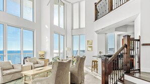 Amplia sala de estar con techo alto, ventanas grandes y vista al océano. Se ve una escalera con baranda de madera que conduce a un nivel superior.