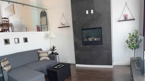 A modern living room with a grey sectional sofa, a black ottoman, a fireplace, and a dining area with a table and chairs.