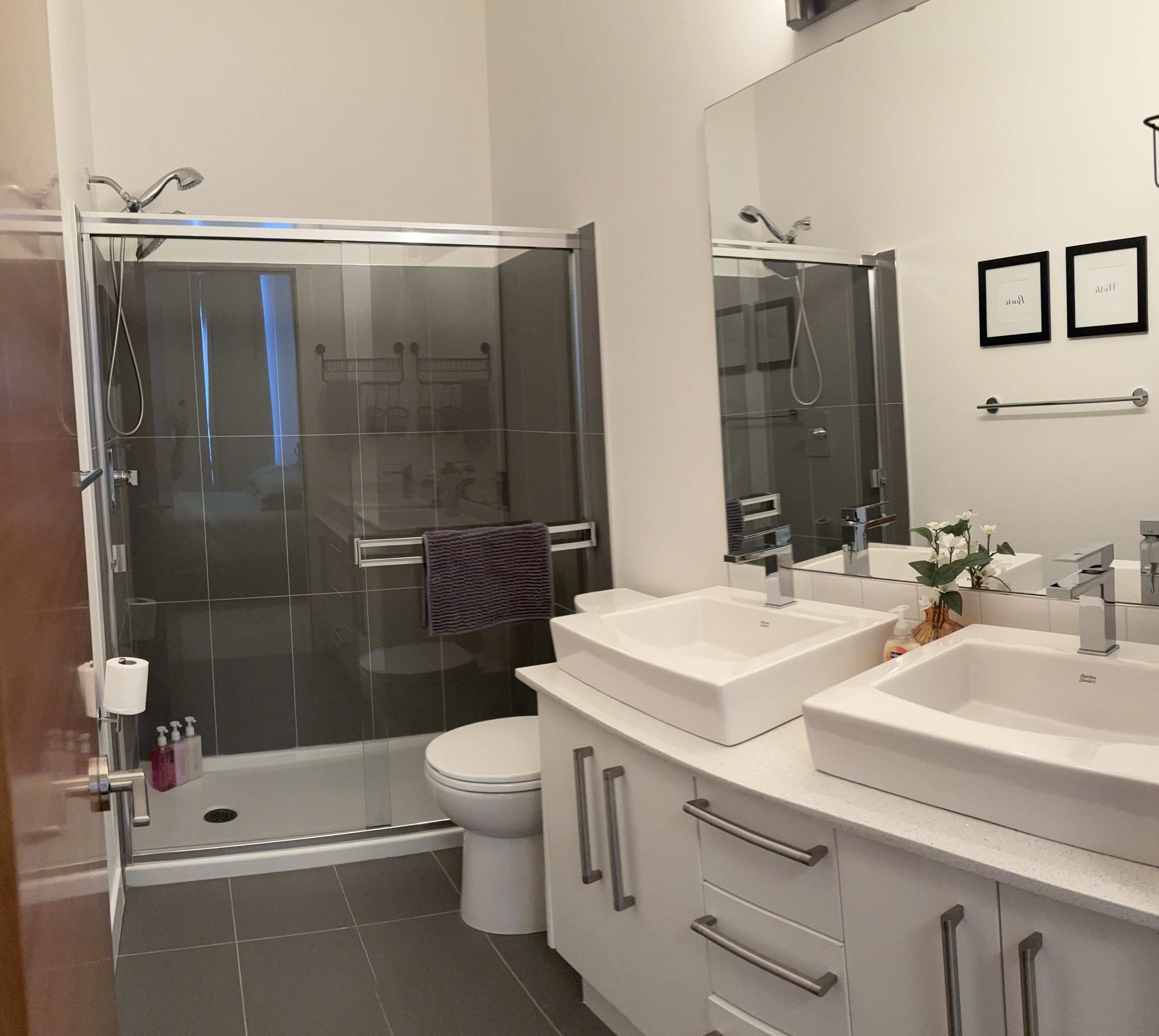 Un baño moderno con ducha envuelta en vidrio, inodoro y dos lavamanos con muebles bajos.