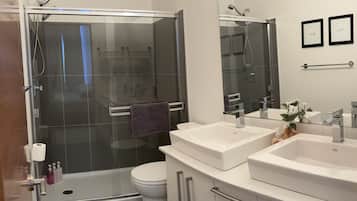 Un baño moderno con ducha envuelta en vidrio, inodoro y dos lavamanos con muebles bajos.