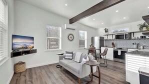 Una sala de estar moderna con un sofá, una mesa de centro y un televisor montado en la pared. Hay una zona de cocina con microondas, horno y fregadero.