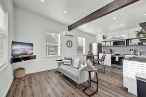 A modern living room with a grey sofa, a coffee table, and a TV mounted on the wall. There is a kitchen area with a microwave, stove, and cabinets.