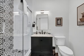 A modern bathroom with a black cabinet, white sink, and toilet. There is a shower curtain with a black and white pattern, and a framed picture on the wall.