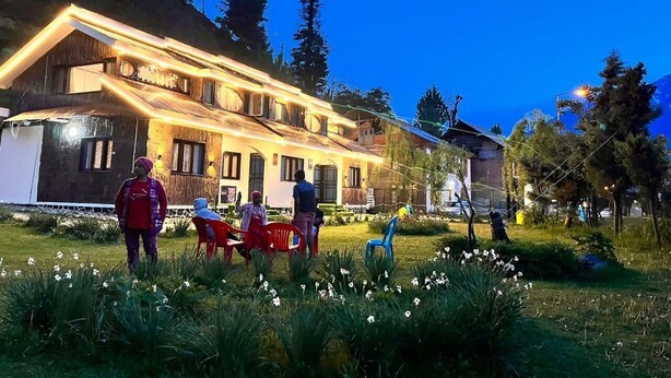 A building with a sloped roof and multiple windows, surrounded by a well-maintained garden with white flowers and green grass. - WATERFRONT PINE PAHALGAM (Anantnag)