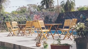 Un área de estar al aire libre con sillas y mesas de madera, un mural en la pared y una piscina al fondo.