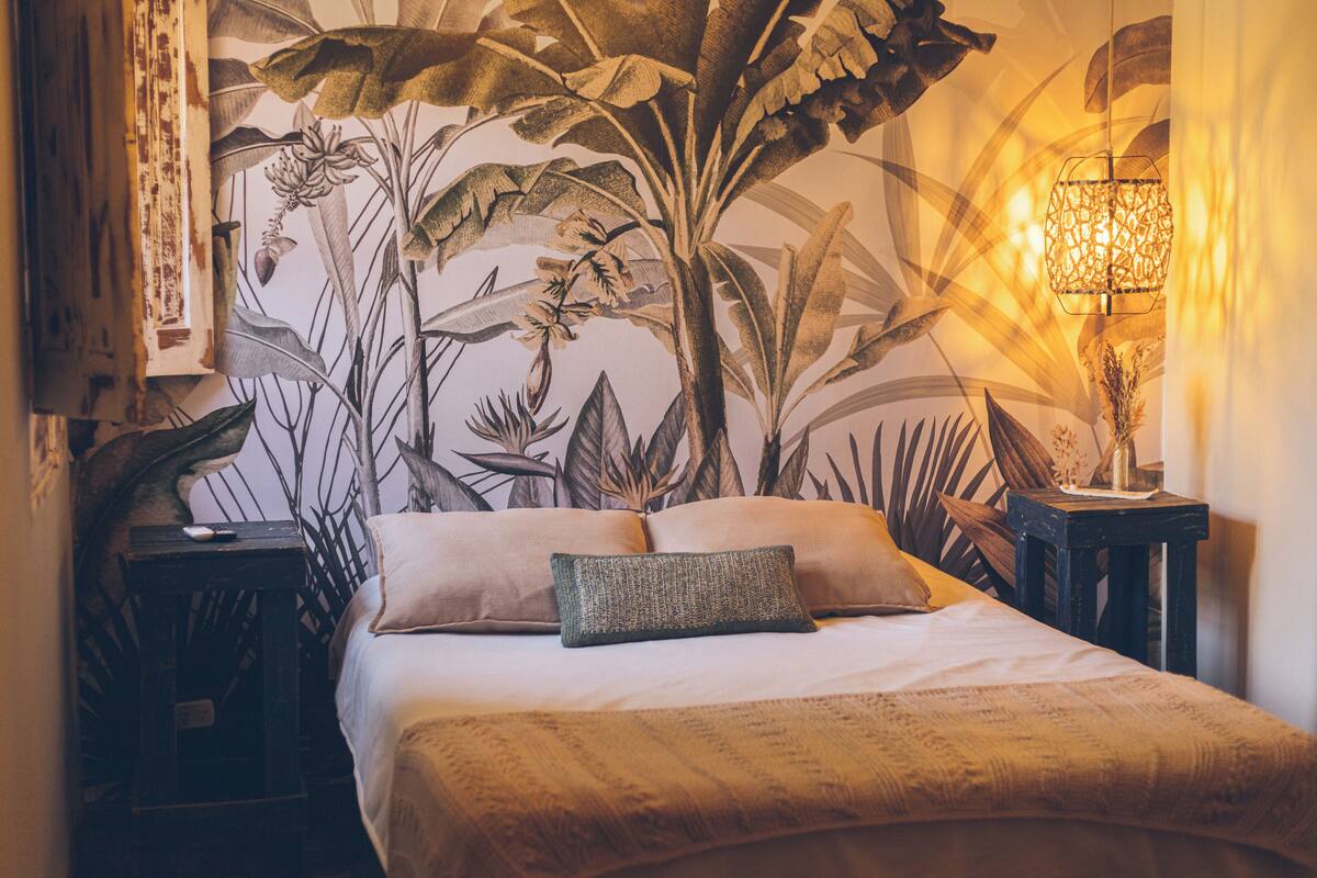 Een slaapkamer met een bed, kussens, een nachtkastje, een lamp en een muurschildering.