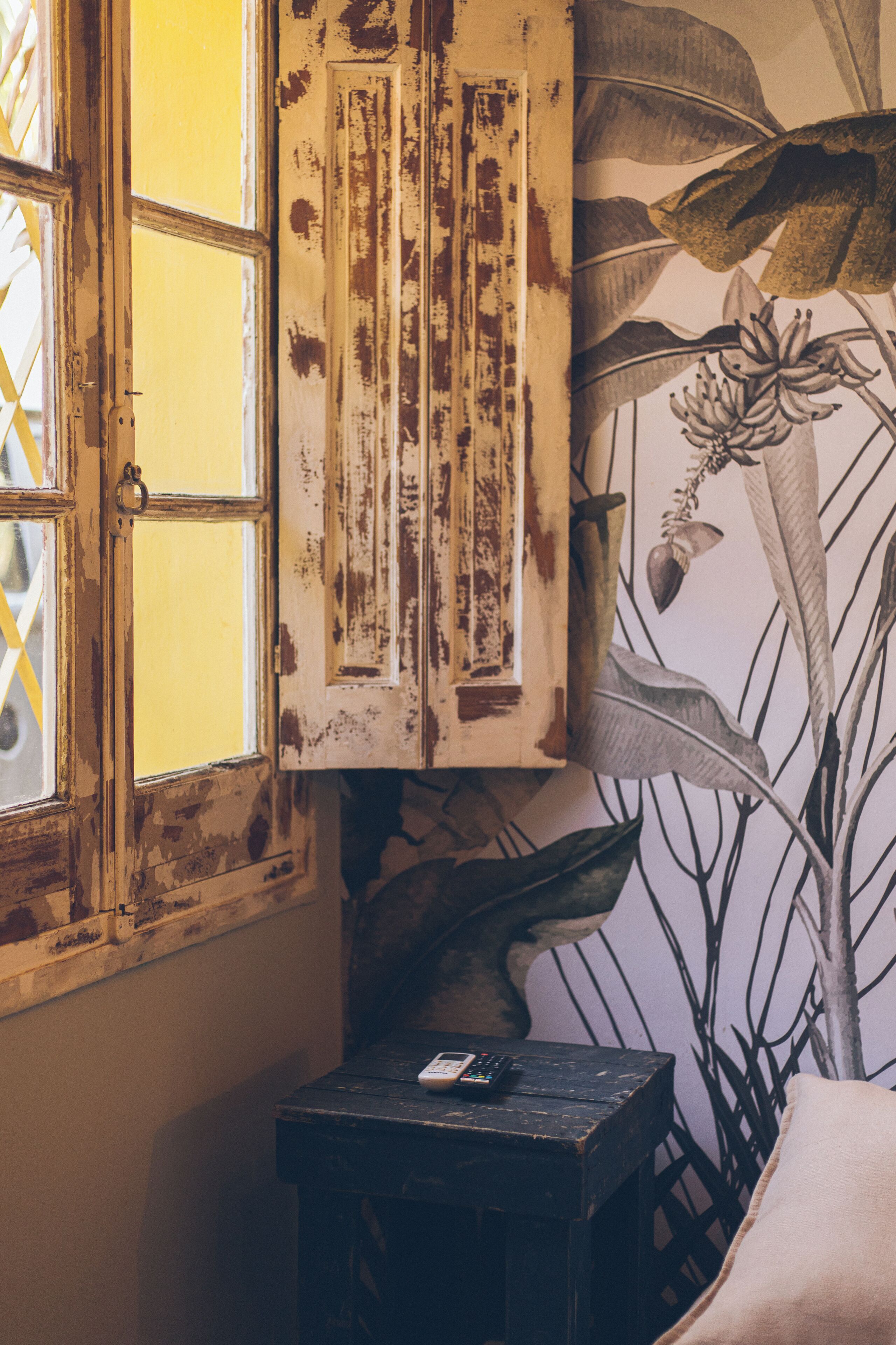 Una habitación con ventana, una pequeña mesa con un control remoto y un mural botánico en la pared.