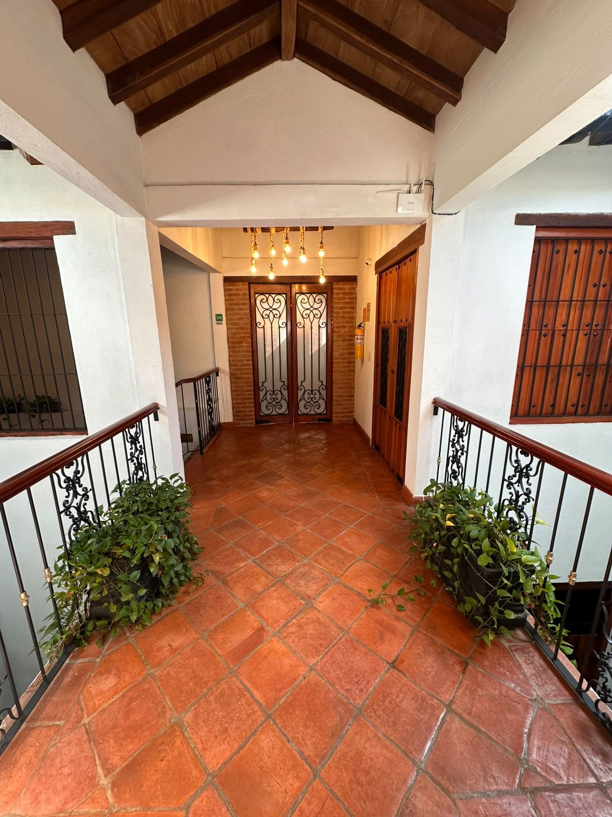 A corridor with a tiled floor, wooden ceiling beams, and a double door with decorative ironwork.