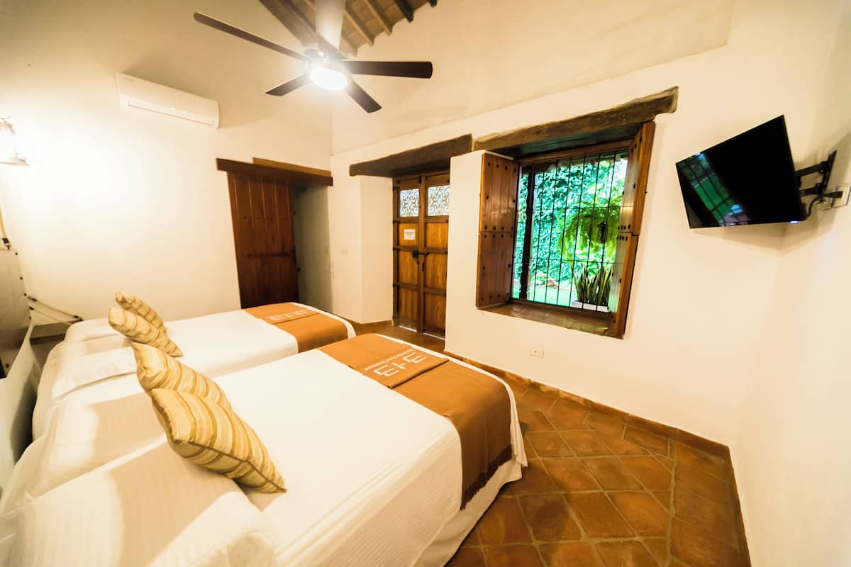 A hotel room with two beds, a ceiling fan, a TV mounted on the wall, and a window with a view of greenery.