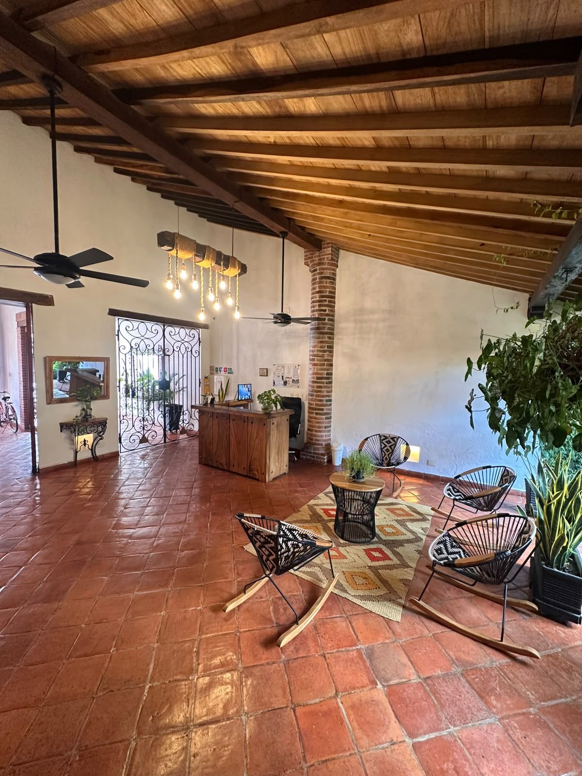 A spacious indoor area with a high ceiling, wooden beams, and a tiled floor. There are several chairs and a table, a reception desk, and a large window with decorative ironwork.